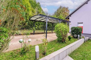 A covered outdoor area with a metal roof, surrounded by greenery and a building with a red door.