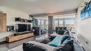 Ett modernt vardagsrum med en stor platt-TV, en soffa, ett soffbord och utsikt över naturen genom stora fönster.