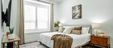 Un dormitorio con cama, ventilador de techo, ventana con cortinas, mesita de noche con lámpara y televisor montado en la pared.