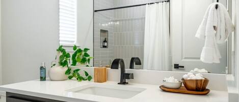 Ein modernes Badezimmer mit einem weißen Waschbecken, einem schwarzen Wasserhahn und einem großen Spiegel. Auf dem Waschtisch stehen ein Topfpflanze, eine hölzerne Seifenschale und eine Schale mit Wattebäuschen.