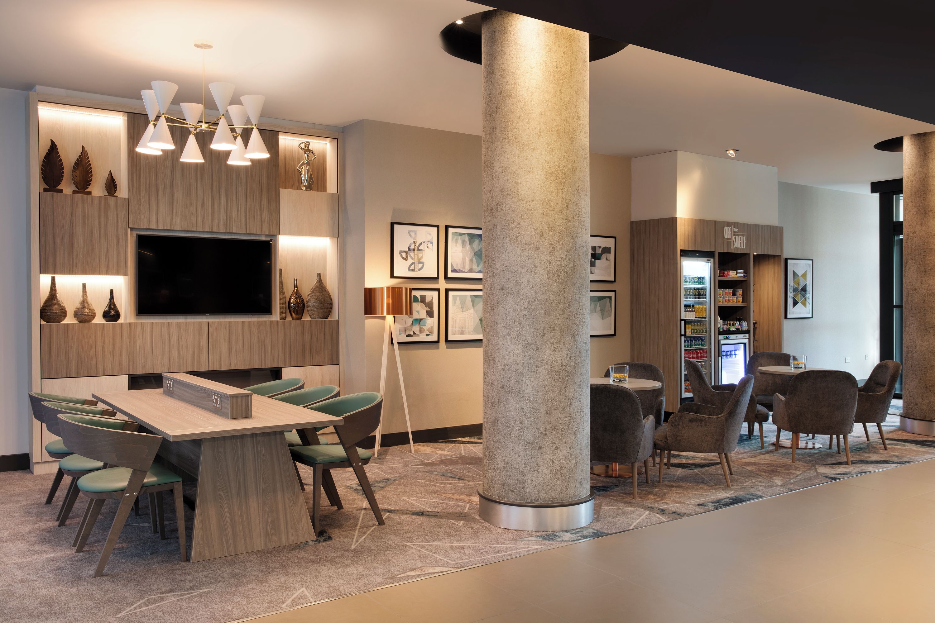 Un moderno hall de hotel con zona de comedor, un pilar grande y un refrigerador con aperitivos.