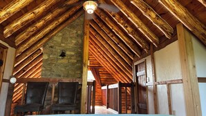 A rustic room with a sloped wooden ceiling, a stone wall, and a ceiling fan. There are two chairs and a bed with a blue bedspread. - Chalet-Style Log Home  – Waynesville, Ohio (Waynesville)