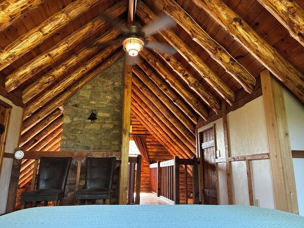 A rustic room with a sloped wooden ceiling, a stone wall, and a ceiling fan. There are two chairs and a bed with a blue bedspread.