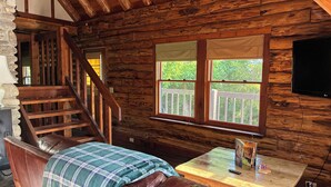 A wooden cabin interior with a wooden staircase, a couch with a plaid blanket, a table with a remote control, and a TV mounted on the wall. - Chalet-Style Log Home  – Waynesville, Ohio (Waynesville)