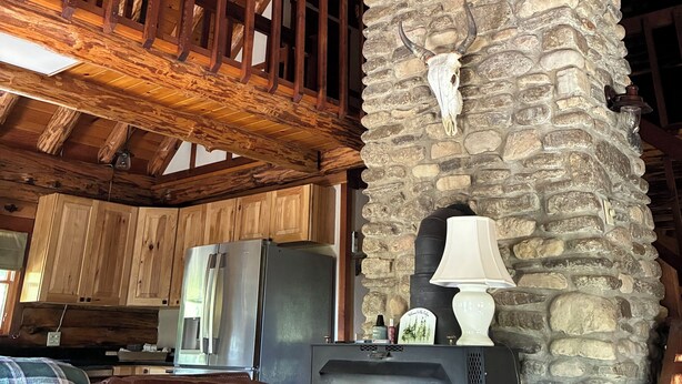A rustic interior with exposed wooden beams, a stone fireplace with a cow skull, and a vintage stove. - Chalet-Style Log Home  – Waynesville, Ohio (Waynesville)