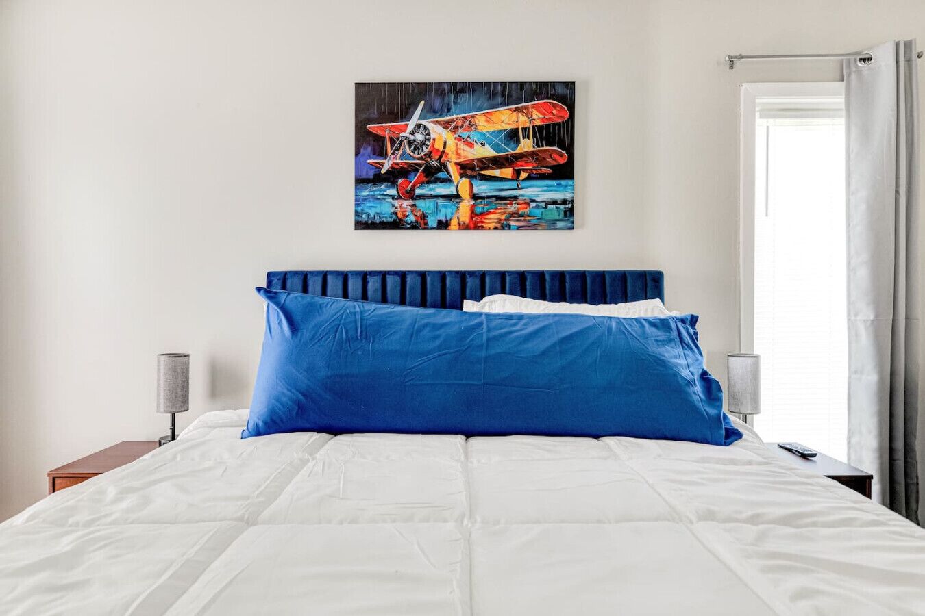Una cama con ropa de cama blanca y almohadas azules, un cabecero, un cuadro de un avión y una mesita de noche con una lámpara.