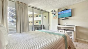A hotel room with a bed, a TV, a desk, and a balcony with a view of a building with stairs.