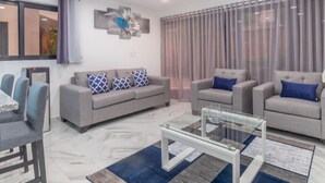 A modern living room with a grey sofa, blue and white patterned cushions, a glass coffee table, and a large window with curtains. - Ala Wishes Luxury Condo (Sosúa)