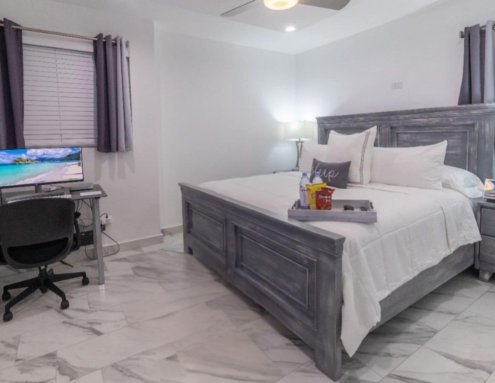 Une chambre à coucher avec un lit, un bureau avec un ordinateur et un téléviseur diffusant une scène de plage.