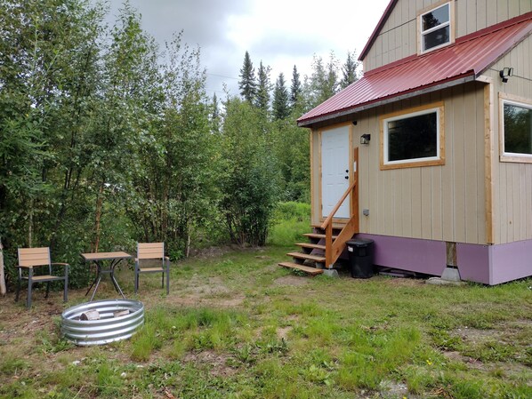 A small wooden cabin with a red roof, a front door, and a set of stairs leading to a porch. There are two chairs and a table outside.