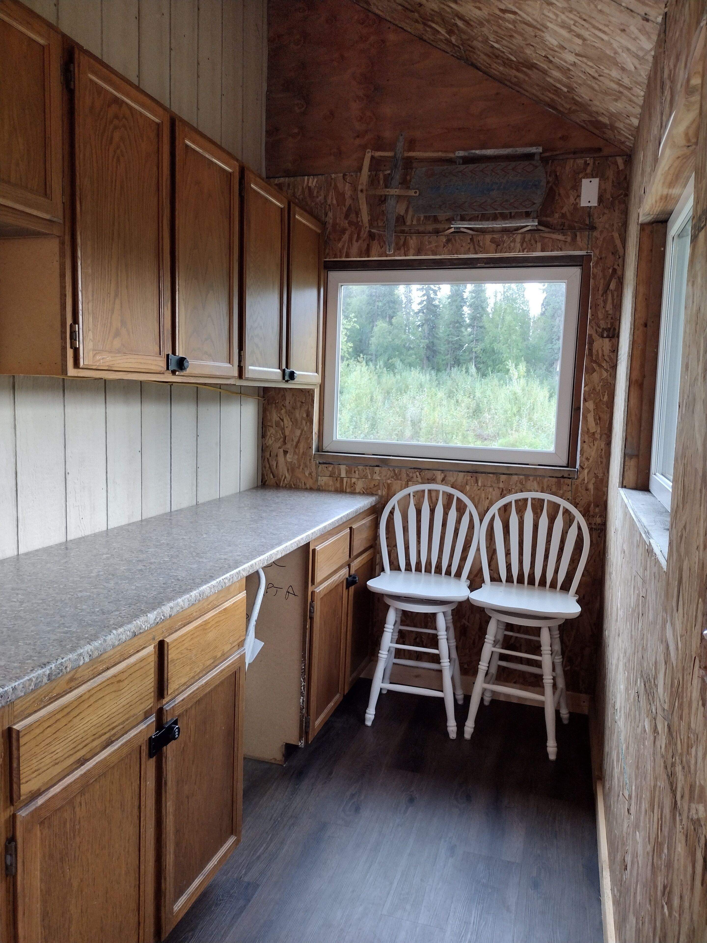 A compact kitchen with wooden cabinets, a granite countertop, two white chairs, and a window overlooking greenery.