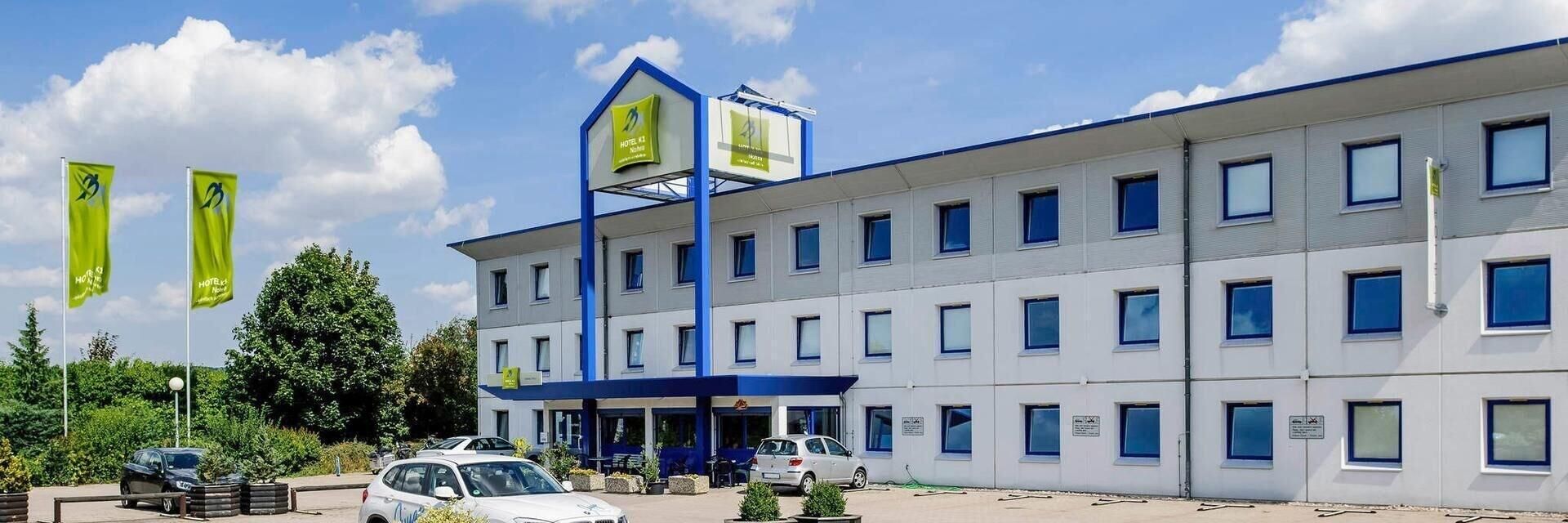 Ein modernes Hotelgebäude mit einem zentralen Turm, zahlreichen Fenstern und einem Parkplatz mit Autos.