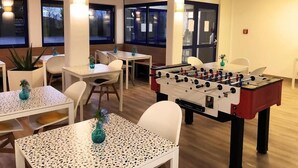 Une pièce avec un baby-foot, plusieurs tables avec des chaises et des plantes en pot.