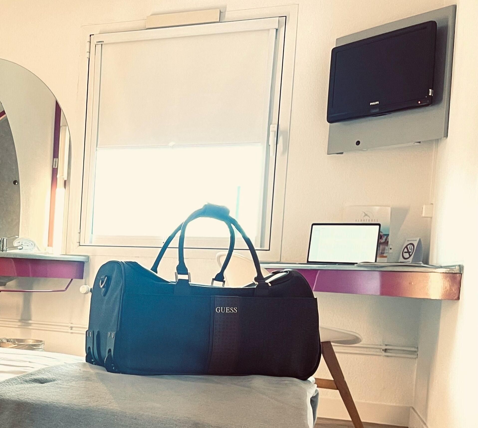 Une chambre d’hôtel avec un lit, un bureau avec un ordinateur portable, une télévision et un sac bleu posé sur le lit.