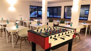 Une pièce comprenant plusieurs tables et chaises, un baby-foot et des fenêtres donnant sur les voitures garées à l’extérieur.