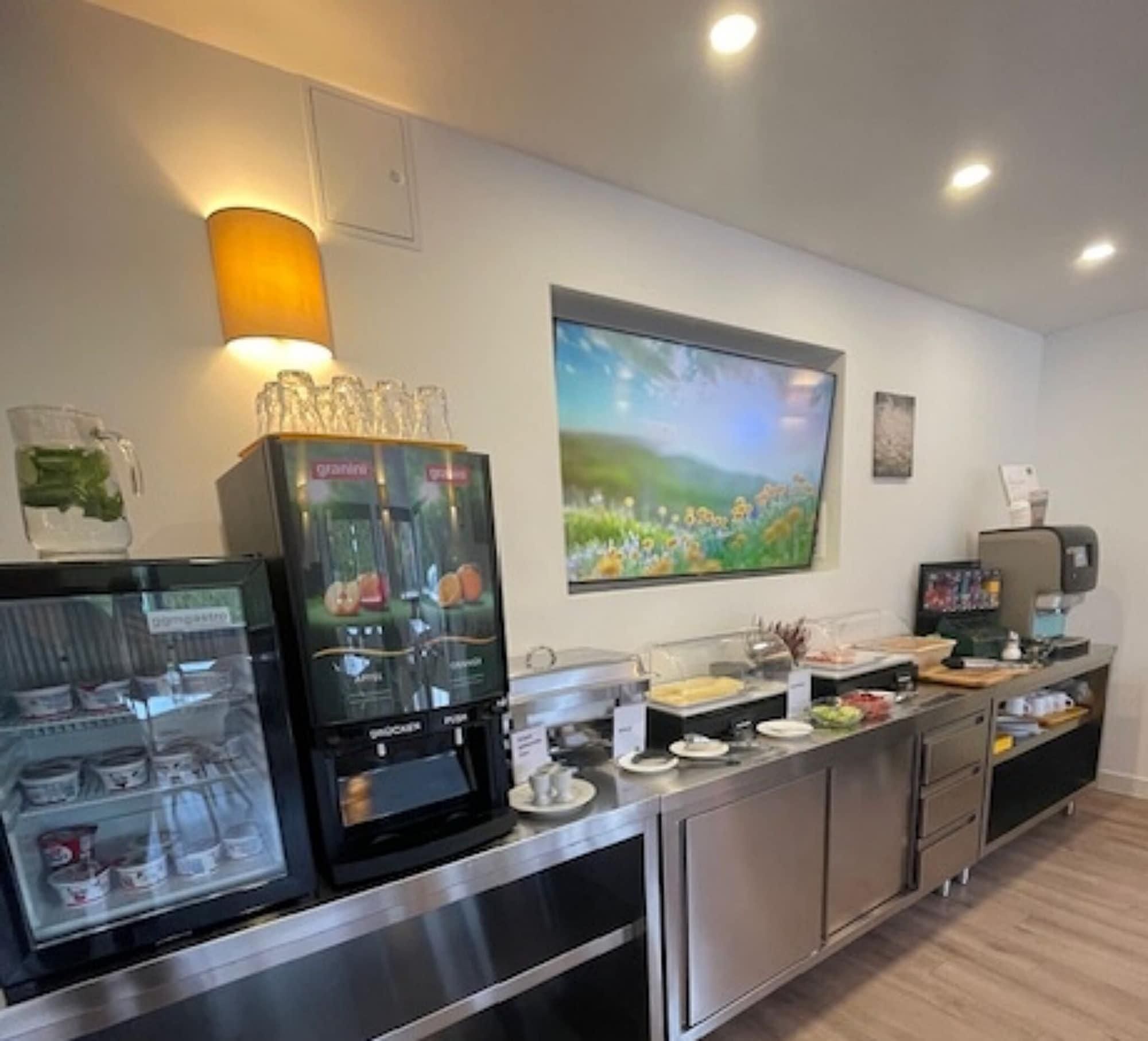 A modern kitchen area with a coffee machine, a glass display case with food items, and a large window with a view of greenery.