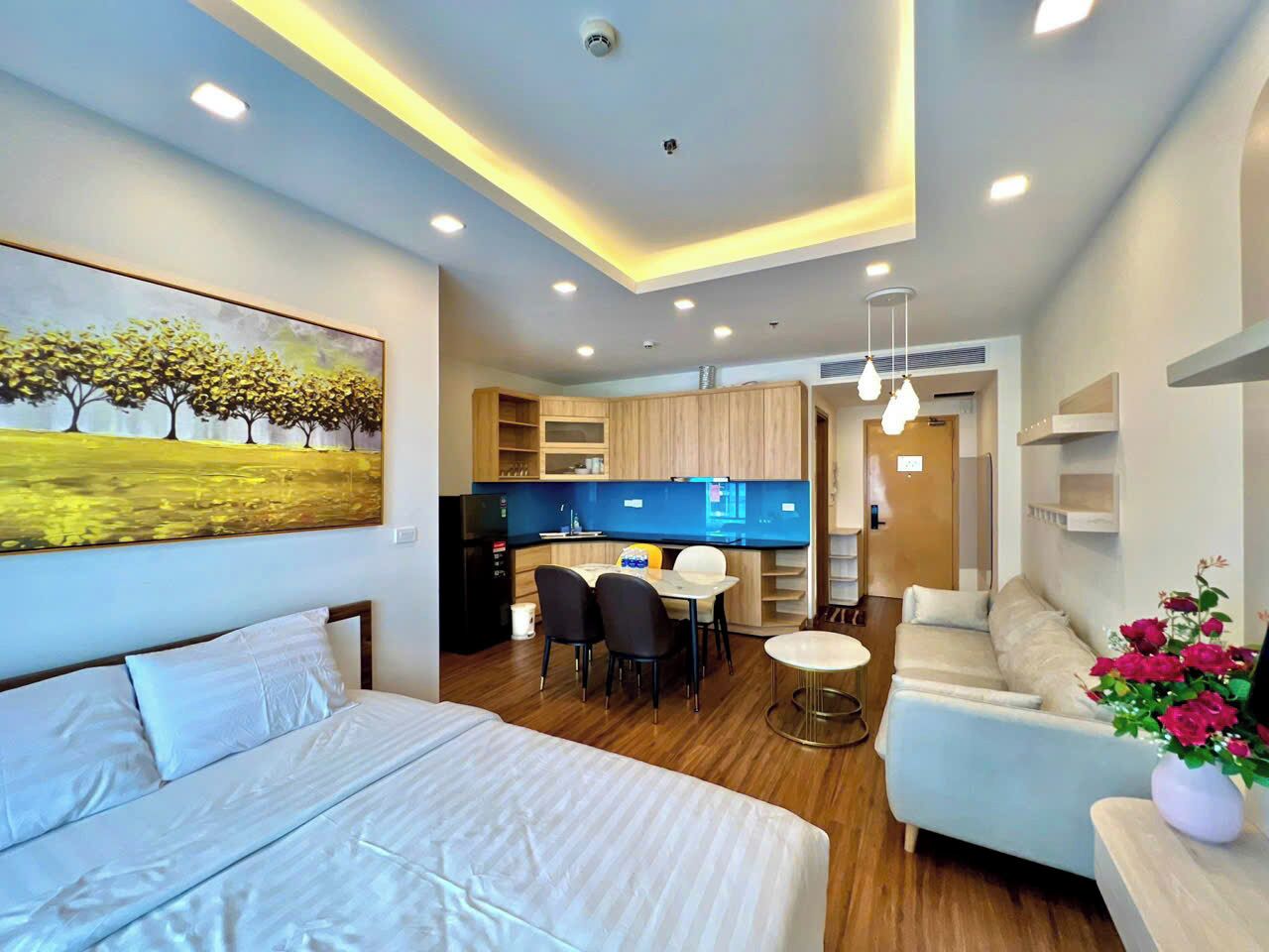 A modern hotel room with a bed, a dining area, a kitchenette, and a living area with a sofa and a TV.