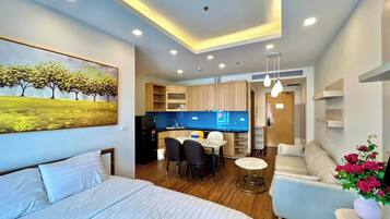 A modern hotel room with a bed, a dining area, a kitchenette, and a living area with a sofa and a TV.
