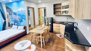 A modern, well-lit room with a wooden floor, a dining area with a table and chairs, a kitchenette with a black countertop, and a mural of a coastal scene on the wall. - FLC Sea Tower Quy Nhon Apartment (Quy Nhon Nam)
