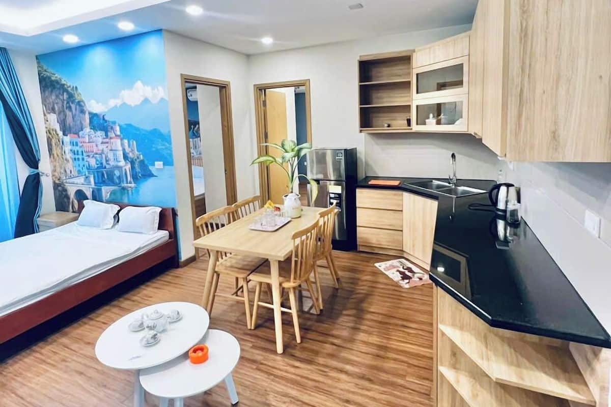 A modern, well-lit room with a wooden floor, a dining area with a table and chairs, a kitchenette with a black countertop, and a mural of a coastal scene on the wall.