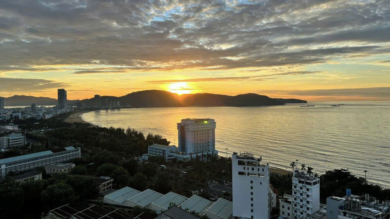 A coastal cityscape at sunset with a prominent building in the foreground, a body of water, and distant hills.