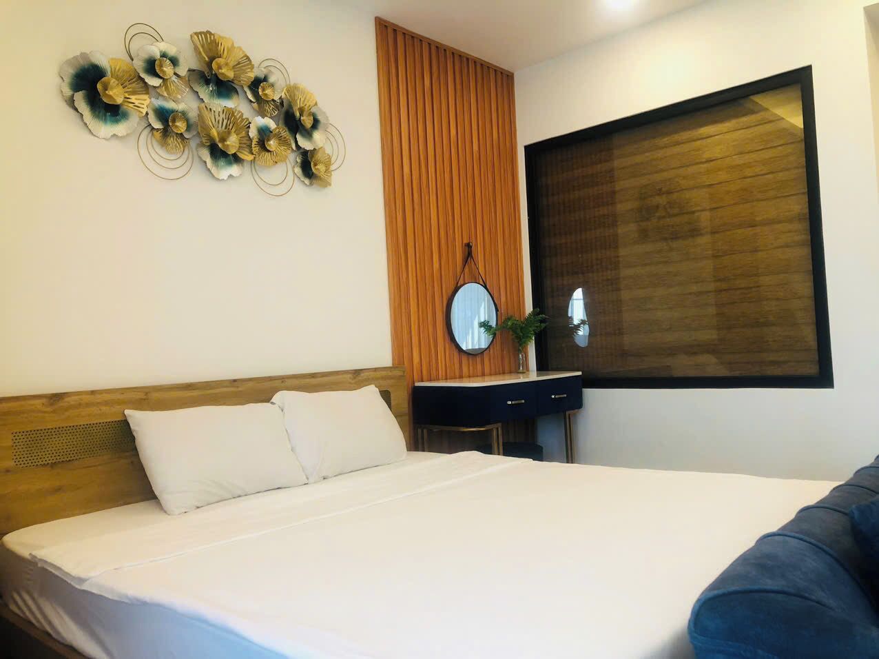 A hotel room with a large bed, a wooden headboard, a wooden panel wall, a round mirror, a dark-colored dresser, and a window with blinds.