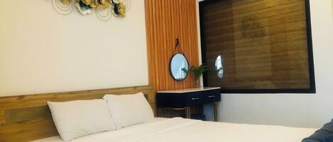 A hotel room with a large bed, a wooden headboard, a wooden panel wall, a round mirror, a dark-colored dresser, and a window with blinds.