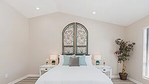 A bedroom with a large bed, two bedside tables, a decorative window, and a potted plant.