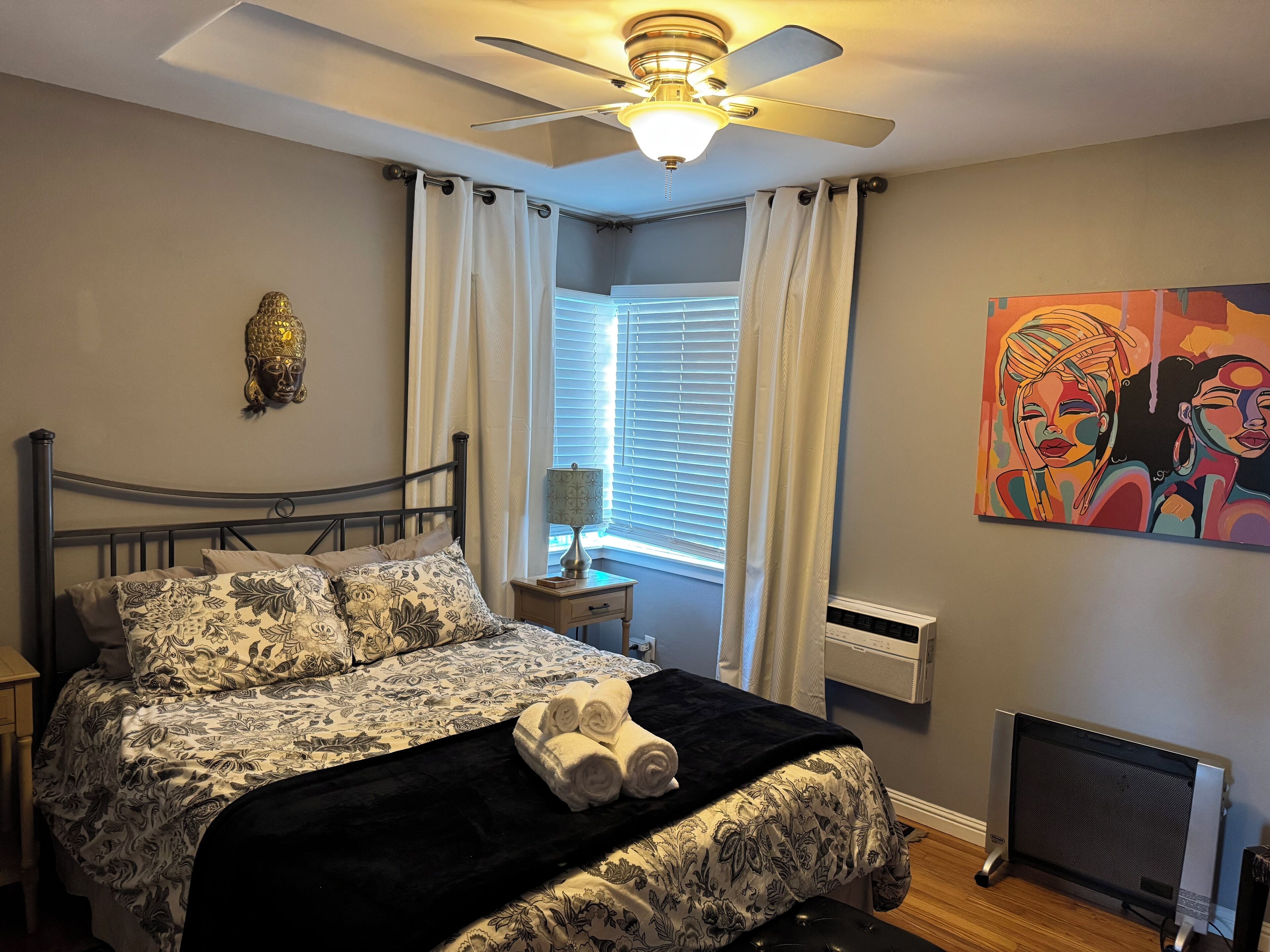Un dormitorio con cama, ventilador de techo, ventana con cortinas, radiador y un cuadro en la pared.