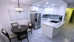 Une cuisine moderne avec des armoires blanches, un réfrigérateur en acier inoxydable, un îlot central doté d’un comptoir en marbre et un coin repas avec une table ronde et des chaises.