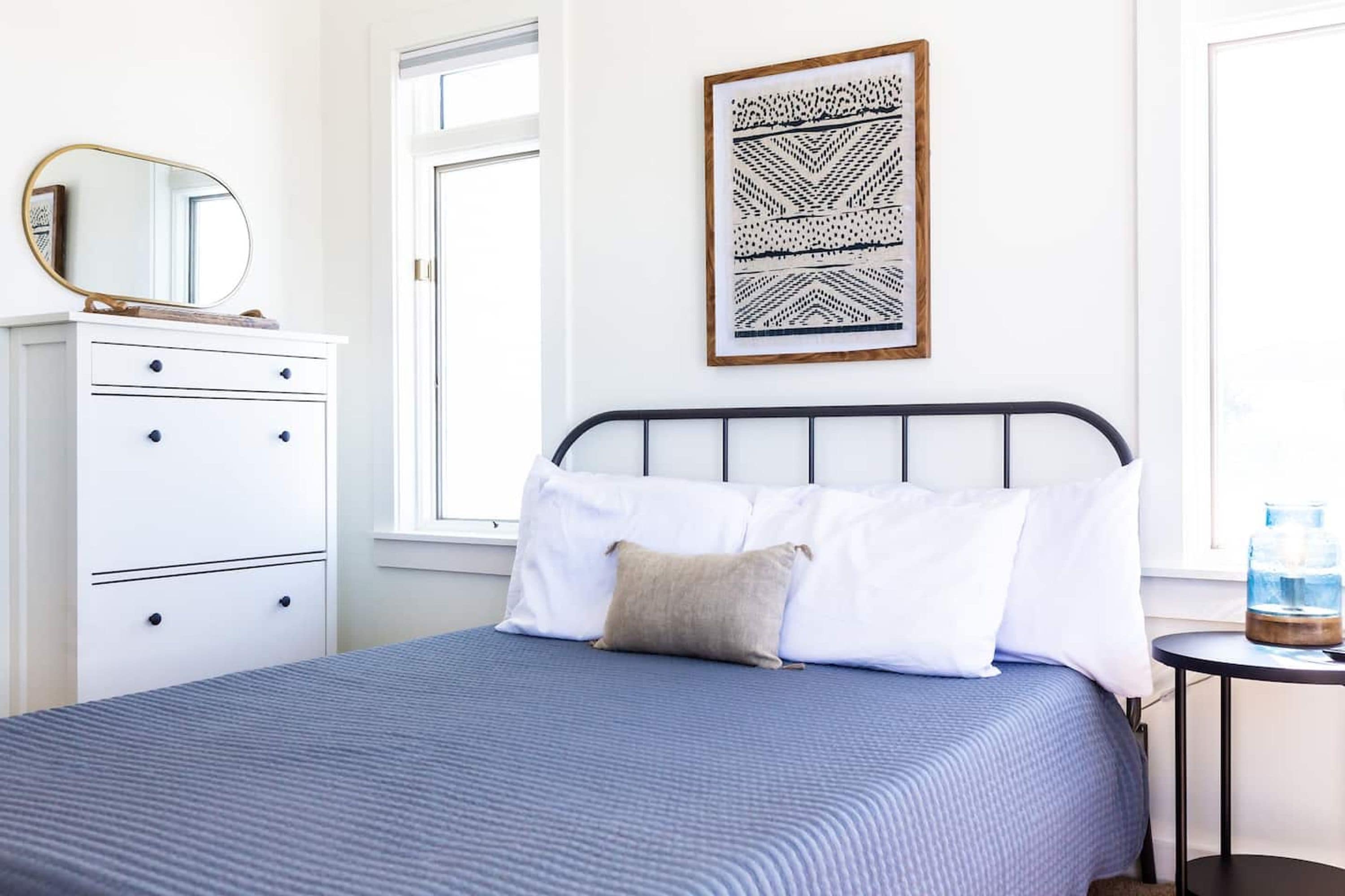 A neatly arranged bedroom with a bed, a dresser, a framed artwork, and a small table with a water bottle.