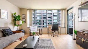 Un salon moderne avec un canapé, une table basse et un bureau avec une chaise. Une grande fenêtre offre une vue sur un immeuble.