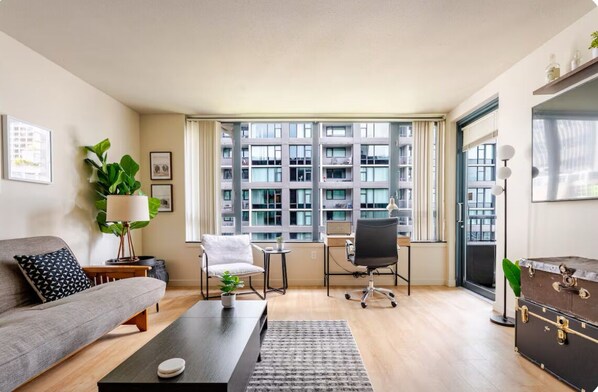 A modern living room with a sofa, coffee table, and a desk with a chair. There is a large window with a view of a building.