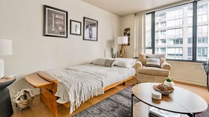 Une chambre moderne avec un lit, un canapé, une petite table et une fenêtre donnant sur des immeubles.