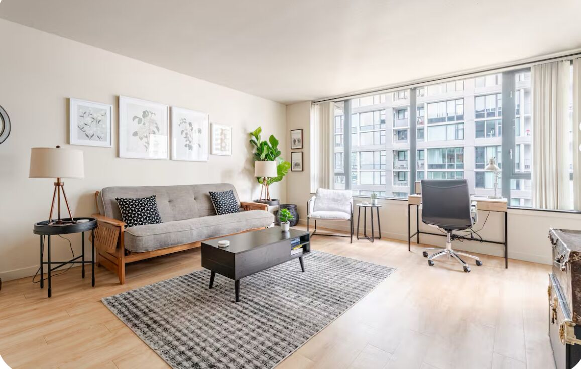 A modern living room with a grey sofa, a coffee table, a side table with a lamp, a white armchair, and a desk with a chair. There are framed pictures on the wall and a large window with a view of buildings.
