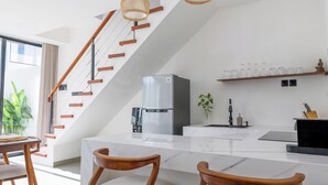 Une cuisine moderne avec un plan de travail en marbre blanc, des chaises en bois, un réfrigérateur en acier inoxydable et un escalier avec des marches en bois et des luminaires suspendus.