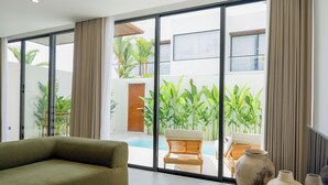 Un salon moderne avec un canapé, une table basse et une vue sur une piscine et un espace extérieur agrémenté de plantes et d’un banc en bois.