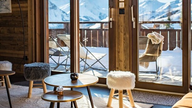 A cozy room with a large sliding glass door leading to a balcony with a mountain view. There are two round tables with stools, a fur-covered ottoman, and a cushioned bench.