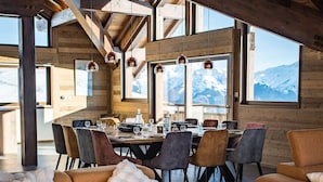 A spacious living area with a large sofa and a dining table set for six. The room features exposed wooden beams and large windows offering a view of the mountains.