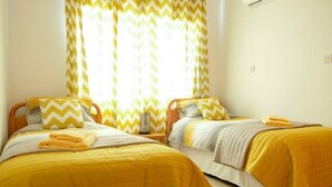 A room with two beds, each with a yellow bedspread and matching pillows. There is a window with yellow and white curtains, and a small table with a lamp between the beds. - A beautiful 2 bedroom apartment with sea views and hot tub .  (Pegeia)