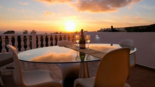 A round glass table with four chairs, a bottle of wine, and glasses on a terrace with a sunset view. - A beautiful 2 bedroom apartment with sea views and hot tub .  (Pegeia)
