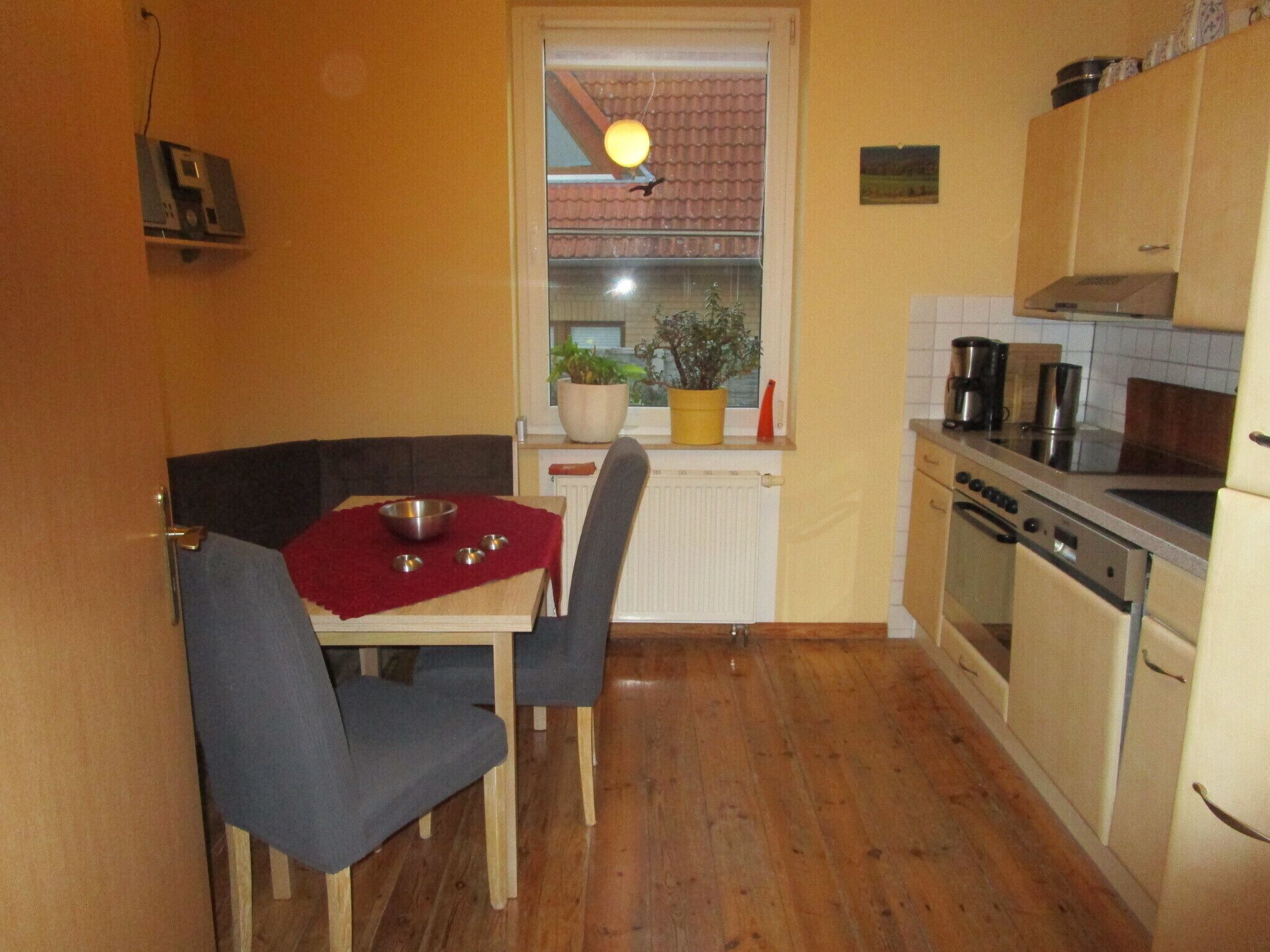 A compact kitchen with wooden flooring, a dining table with two chairs, a built-in oven, and a window with a view of a red-tiled roof.