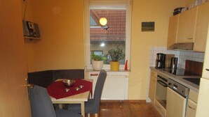 A compact kitchen with wooden flooring, a dining table with two chairs, a built-in oven, and a window with a view of a red-tiled roof. - Vacation apartment near the old town and lake (Neubrandenburg)