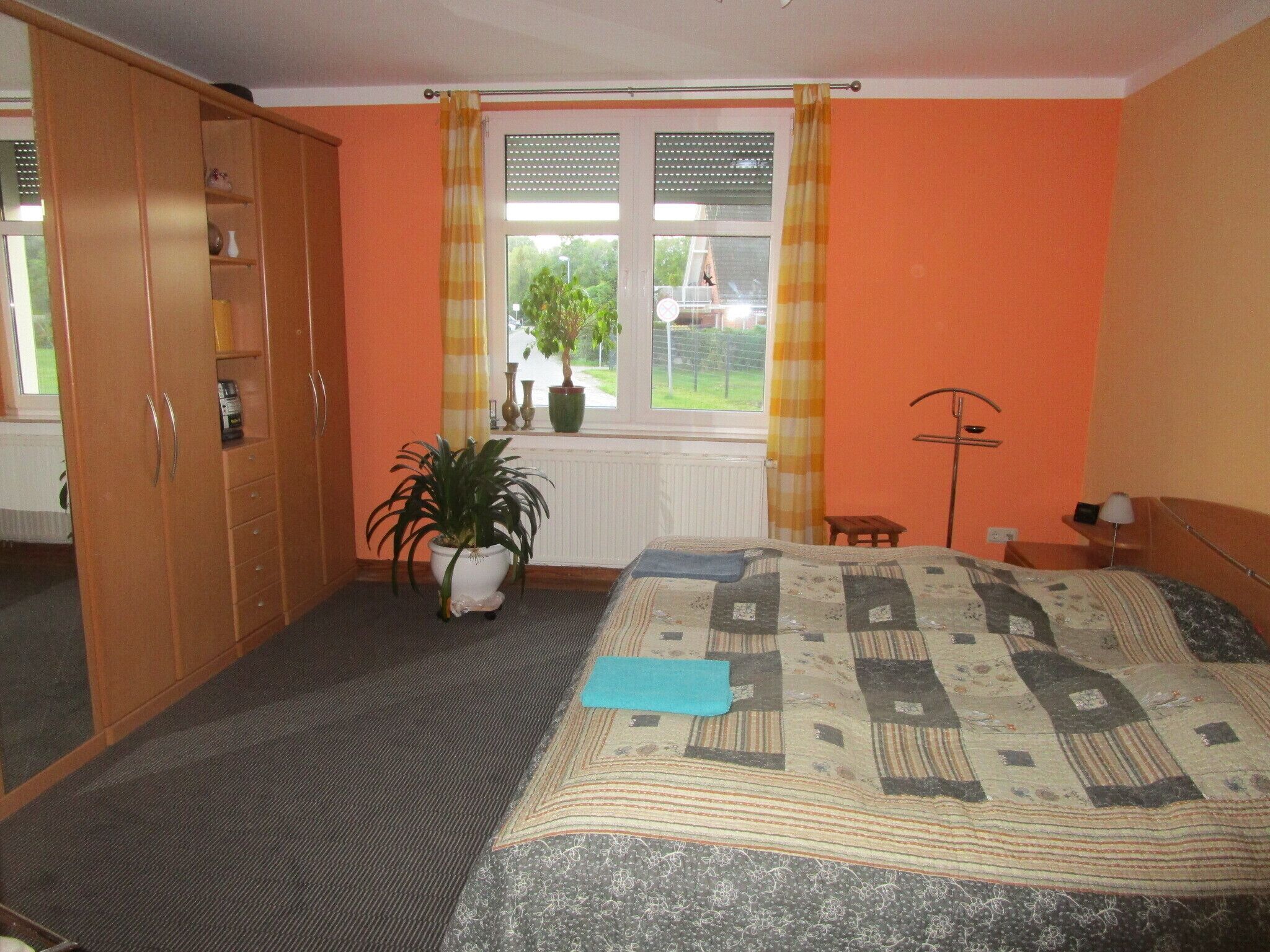 Une chambre avec un mur orange, un lit recouvert d’une courtepointe, une plante en pot, une fenêtre avec des rideaux et une armoire.