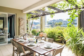 Un comedor con una mesa dispuesta para cuatro personas, plantas de exterior y vistas de edificios al fondo.