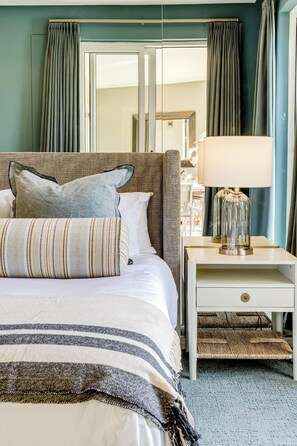 Un dormitorio con cama, almohadas, mesita de noche con lámpara y cortinas.
