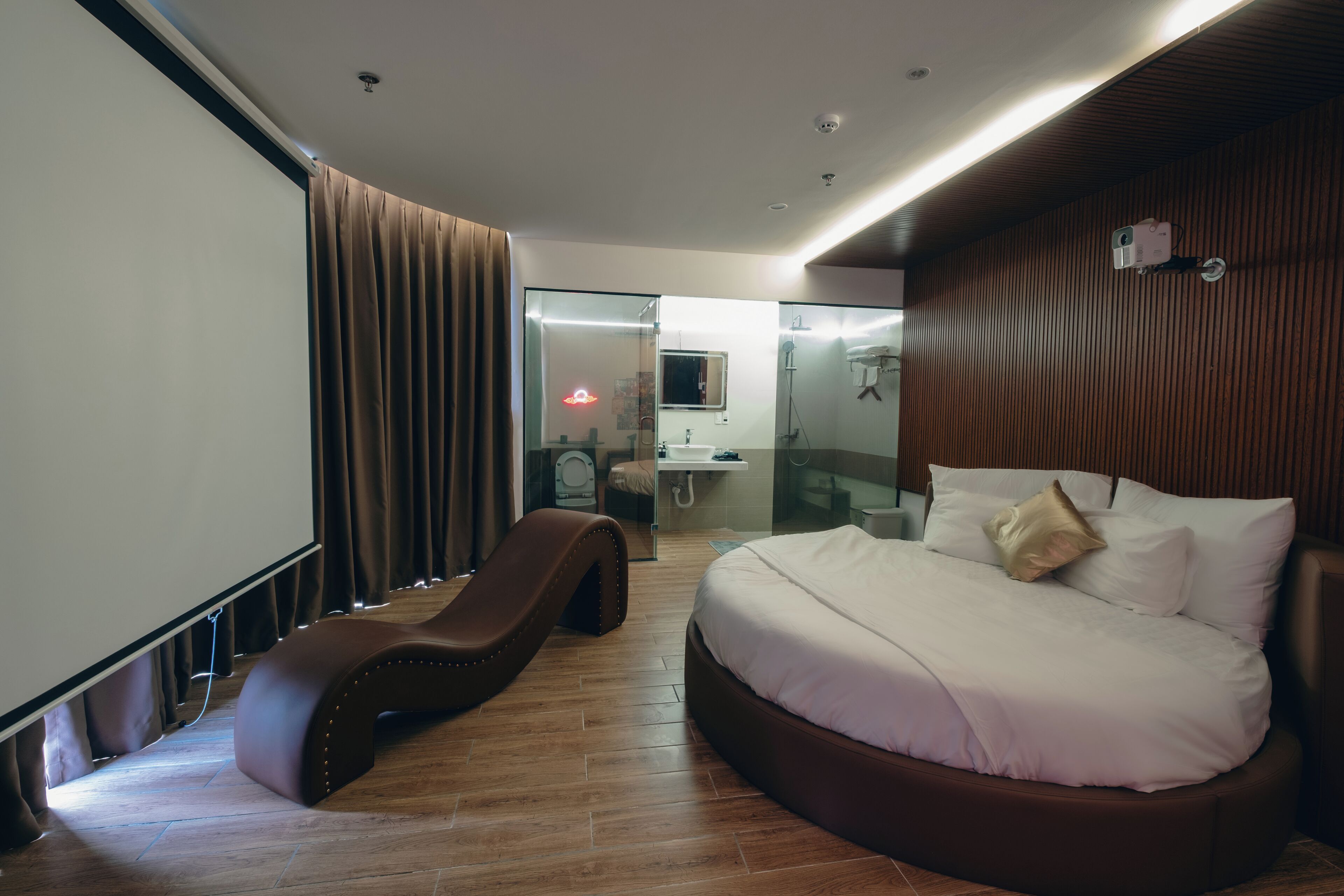 A modern hotel room with a curved bed, a built-in guitar, a glass shower enclosure, and a view of the bathroom.