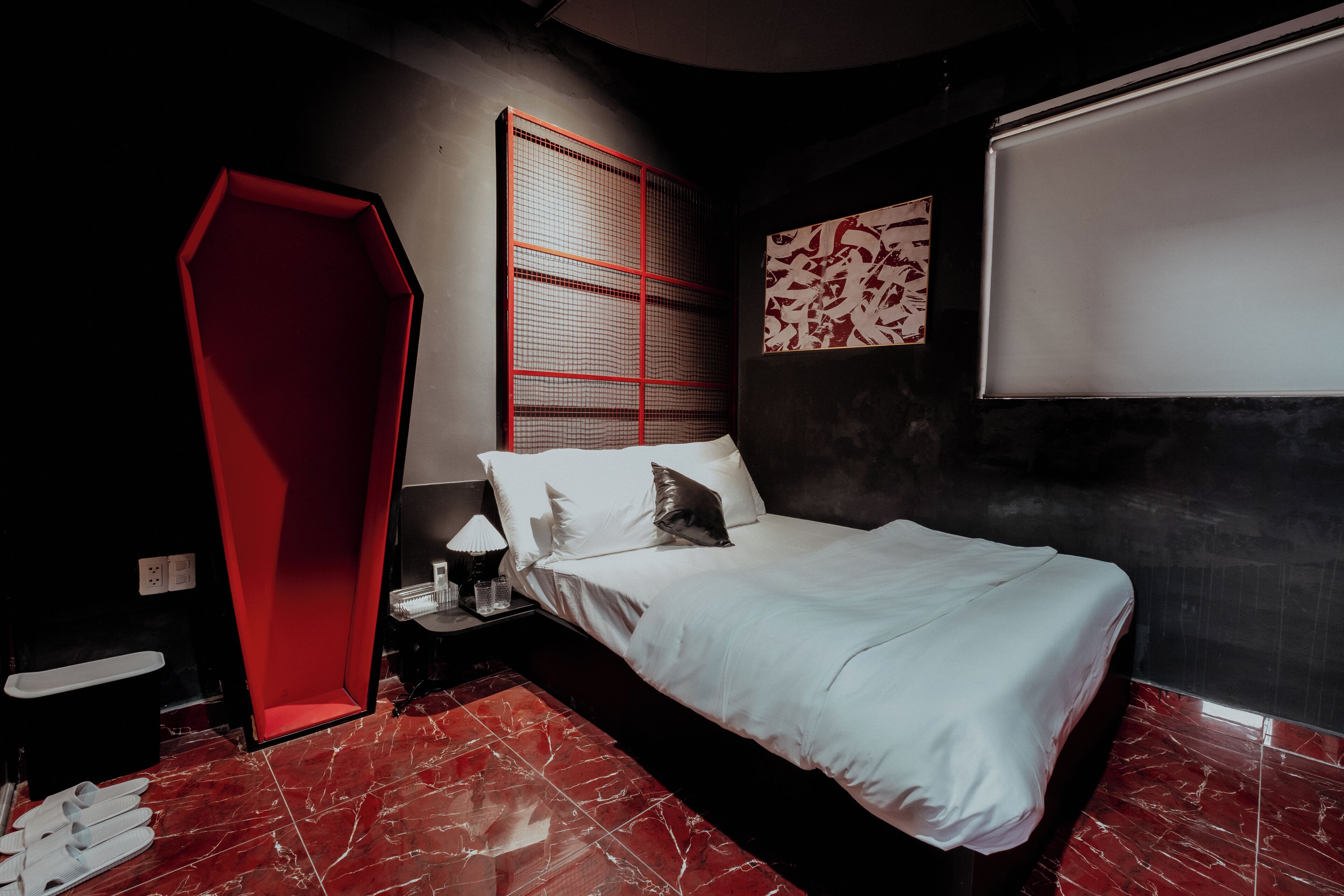 A modern bedroom with a bed, a red accent wall, a large window with blinds, and a whiteboard on the wall.