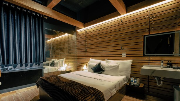 A modern hotel room with a large bed, a bedside table with a lamp, a bathroom with a sink and mirror, and wooden paneling on the walls. - Love in the Atlantis Hotel (Hanoi)
