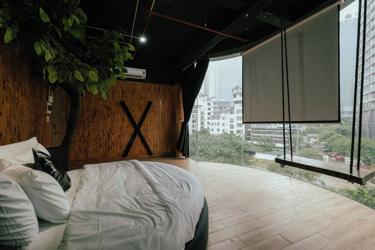 Un dormitorio moderno con una cama grande, una pared de madera, una planta colgante y vistas a un paisaje urbano a través de un ventanal.